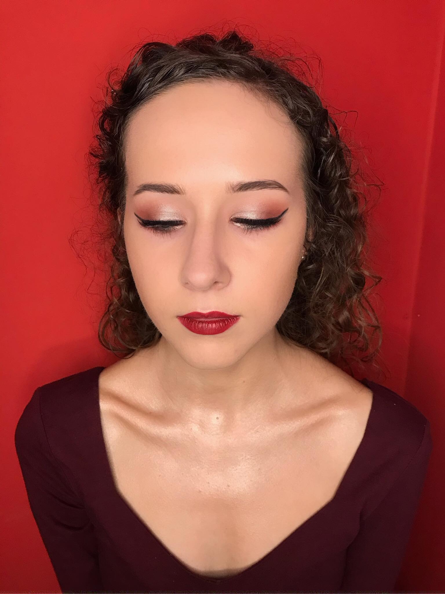 Portret kobiety z zamkniętymi oczami, prezentującej elegancki, wieczorowy look z bordową szminką i kreską na powiekach, na jednolitym czerwonym tle.