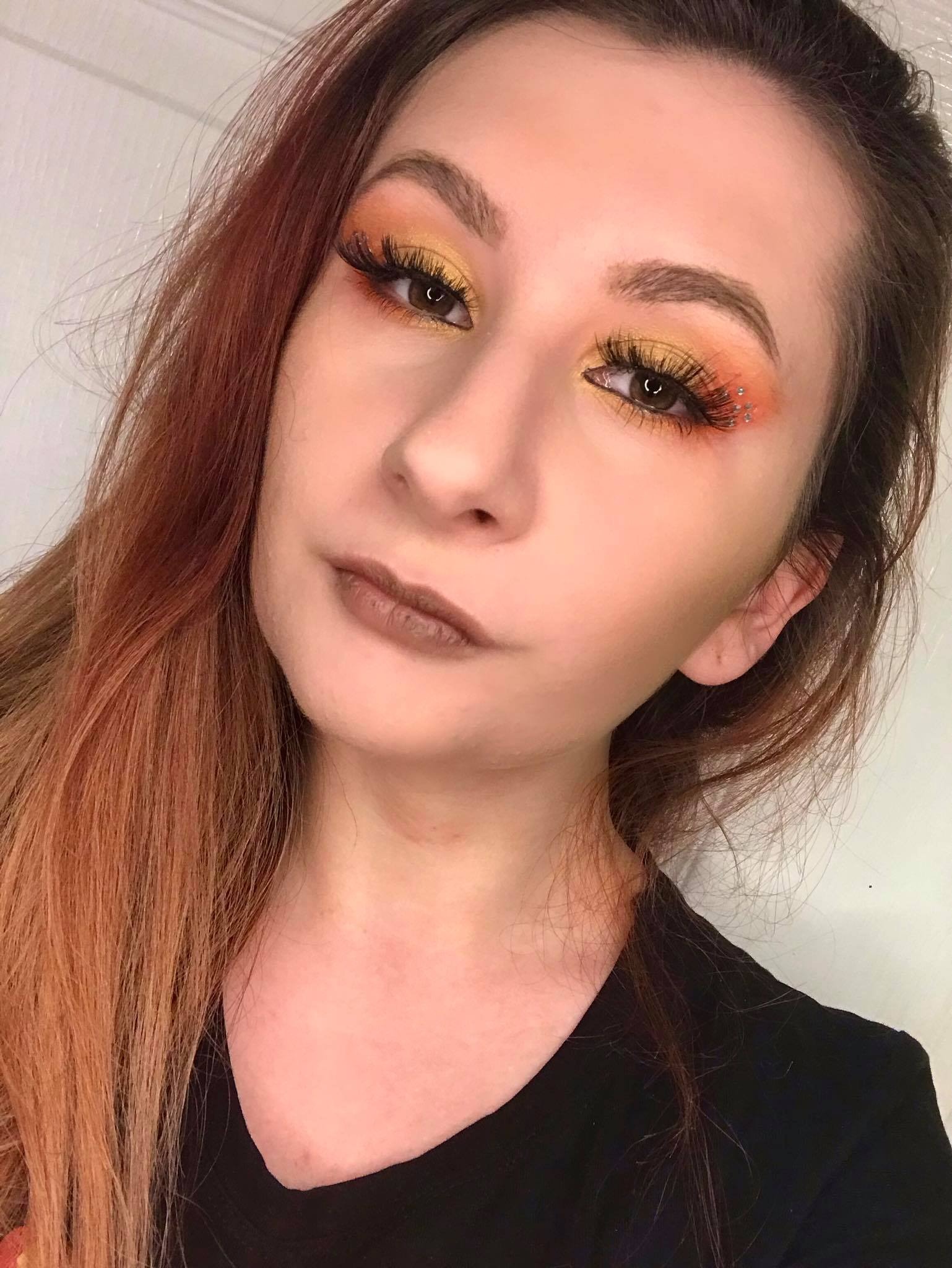 Portret kobiety z pomarańczowo-żółtym cieniowaniem powiek i małymi błyszczącymi aplikacjami przy zewnętrznym kąciku oka, brązowa szminka, włosy w odcieniach rudości.