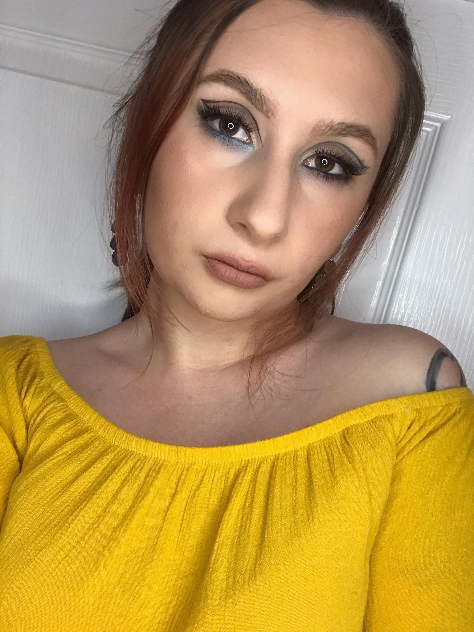Portret kobiety z brązowymi oczami i precyzyjną kreską w kolorze turkusowym na powiekach, w żółtej bluzce z odkrytymi ramionami na tle białych drzwi.