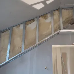 Skośna ściana poddasza w trakcie ocieplania pianą i zabudowy płytami gipsowo-kartonowymi, widoczne metalowe profile, oświetlenie dzienne.