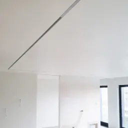 Wnętrze w trakcie wykańczania z zamontowanym oświetleniem liniowym w suficie, widoczne przygotowane otwory w ścianie na instalacje elektryczne i fragment okna.