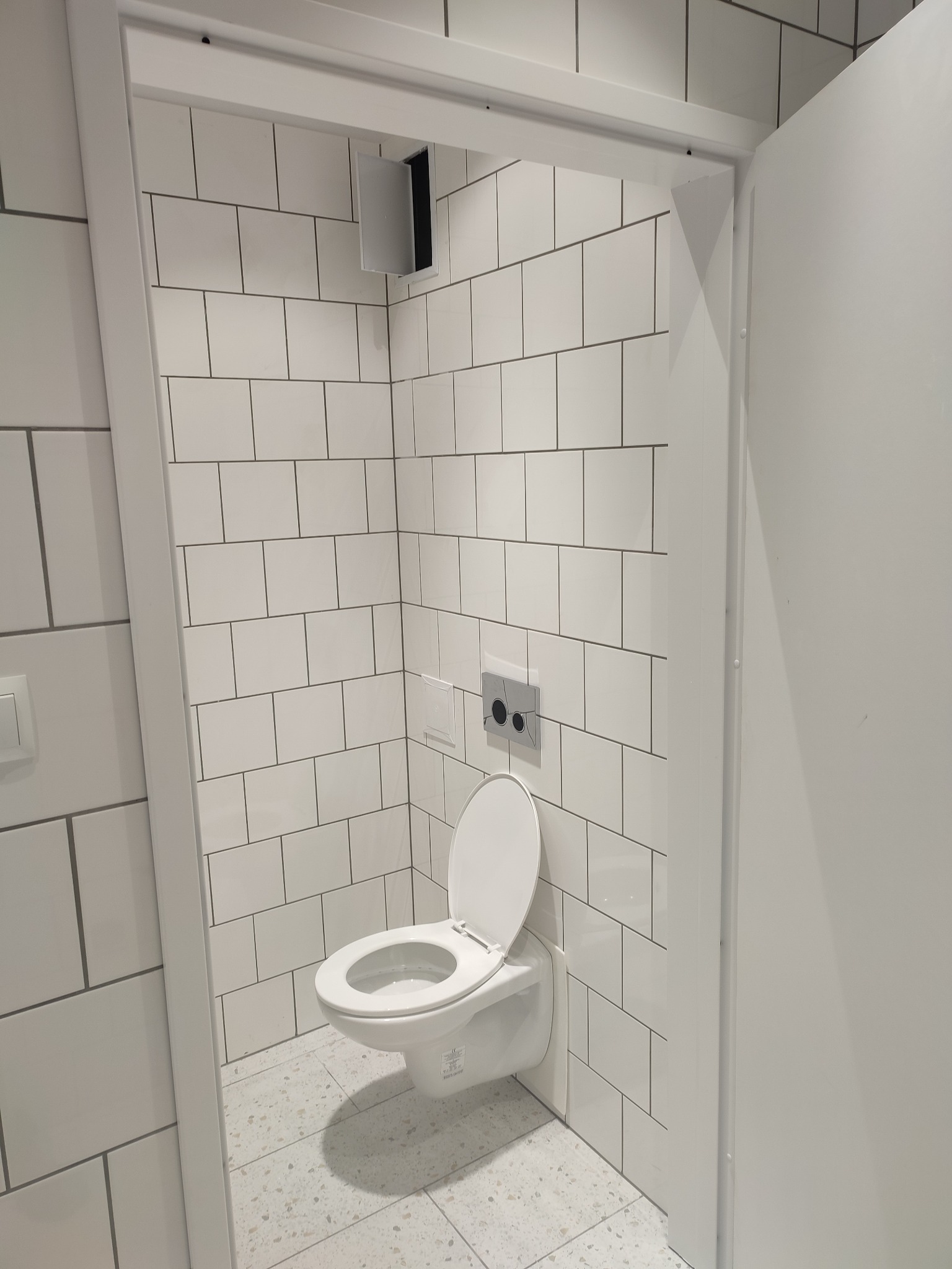 Nowoczesna toaleta z białymi, prostokątnymi płytkami i podtynkową spłuczką, widok z perspektywy wejścia.