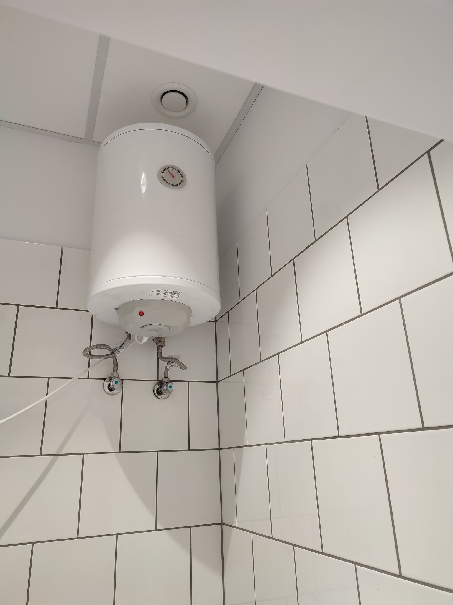 Bojler elektryczny zamontowany na ścianie wyłożonej białymi płytkami w łazience, widoczne zawory doprowadzające wodę i fragment sufitu z kratką wentylacyjną.