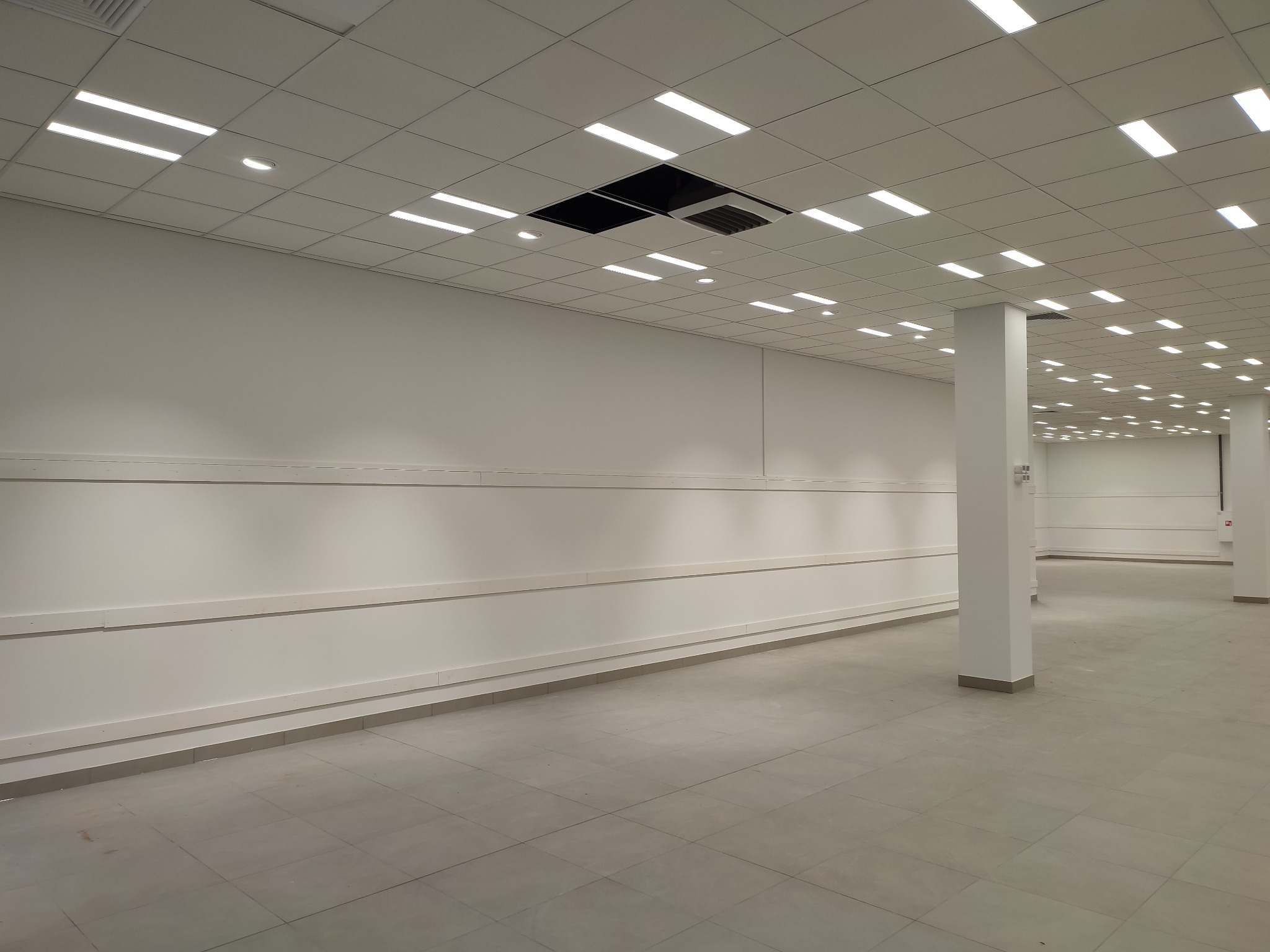 Pusta, jasna przestrzeń biurowa z szarą podłogą z płytek, białymi ścianami z listwami przypodłogowymi i sufitem kasetonowym z oświetleniem LED, widoczne otwory w suficie.