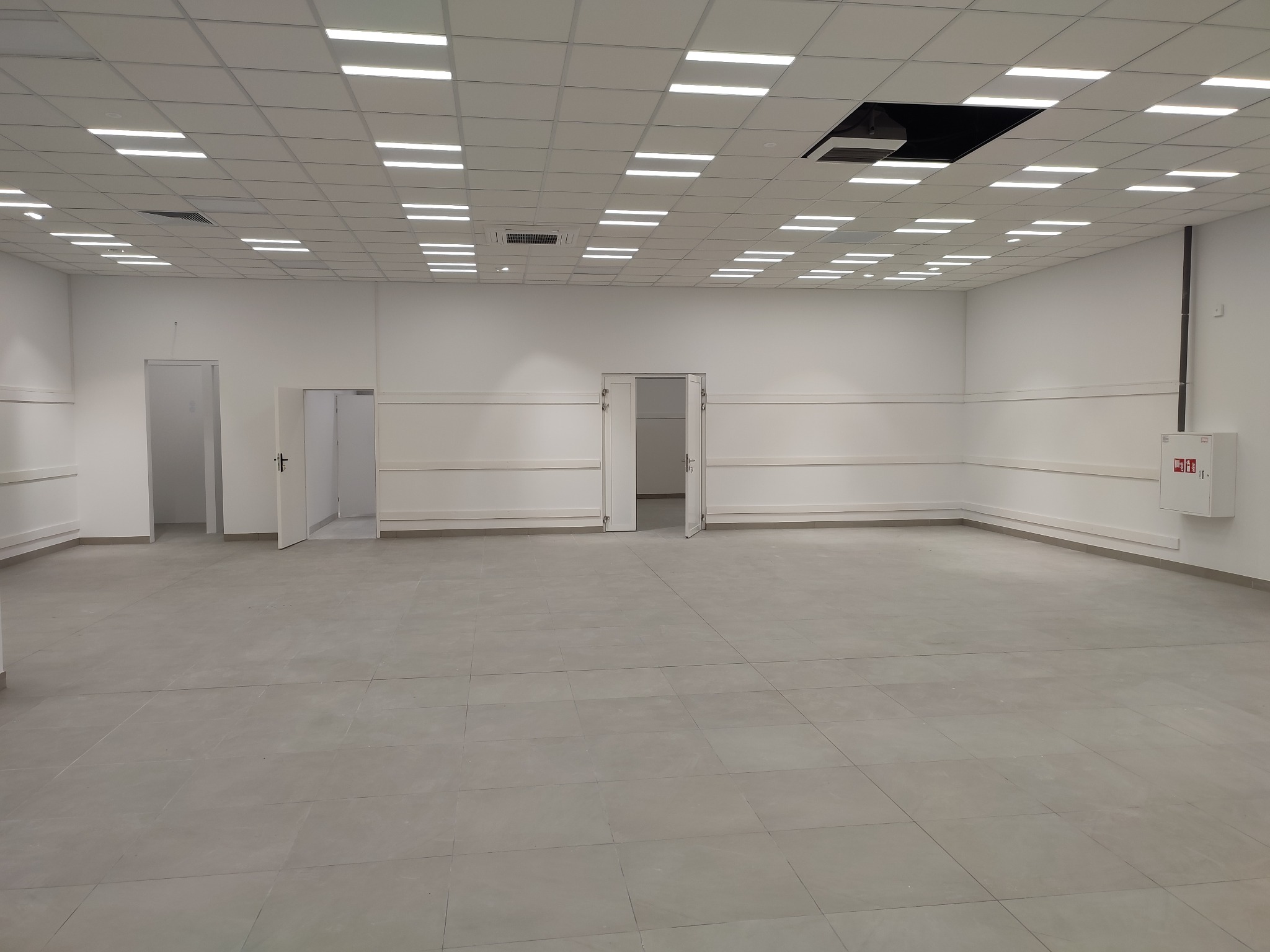 Puste pomieszczenie biurowe z szarą podłogą z płytek, białymi ścianami z listwami przypodłogowymi i sufitem kasetonowym z oświetleniem LED oraz otwartymi drzwiami.