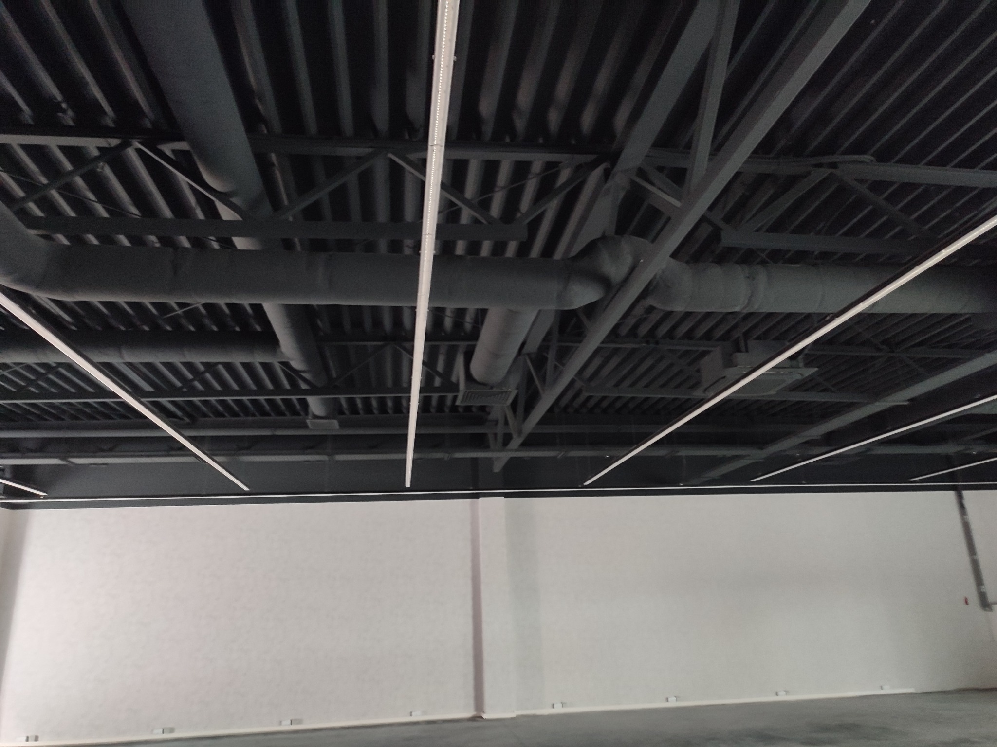 Widok sufitu w pomieszczeniu przemysłowym z widocznymi elementami konstrukcyjnymi, rurami wentylacyjnymi i oświetleniem LED, ściany pomalowane na jasny kolor.