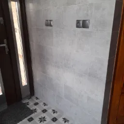 Szara ściana wyłożona podłużnymi płytkami, dwa podwójne gniazdka elektryczne, podłoga z wzorem szarych gwiazd na białym tle, fragmenty drzwi wejściowych.