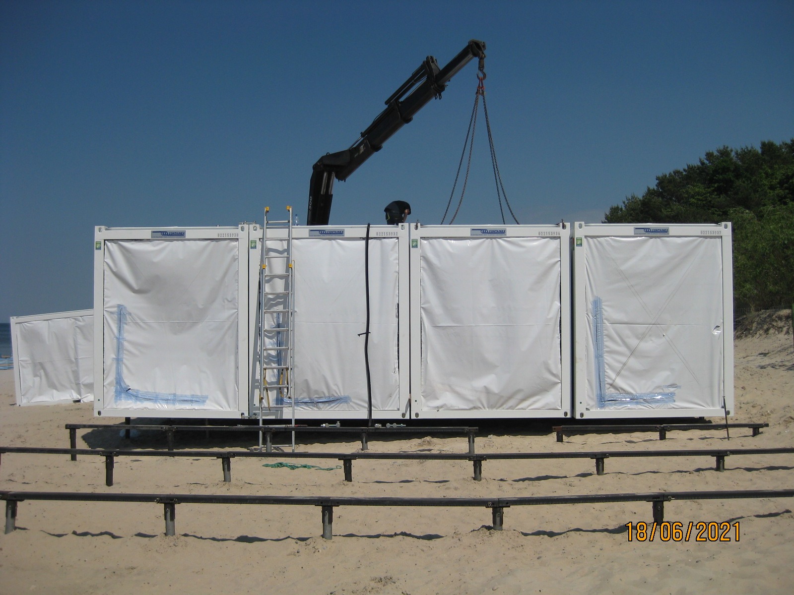 Montaż modułowego pawilonu z kontenerów na plaży, widoczny dźwig podnoszący element, drabina oparta o kontener, w tle las i błękitne niebo.