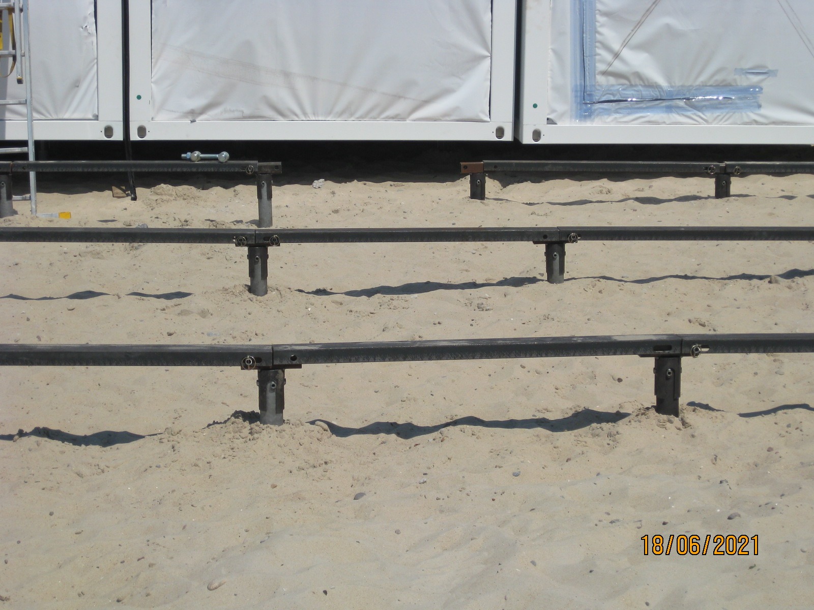 Metalowa konstrukcja nośna na piasku, prawdopodobnie pod budowę modułową, z widocznymi elementami mocującymi i datą 18/06/2021.