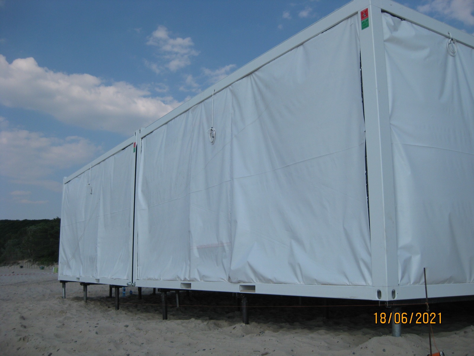 Dwa białe kontenery modułowe, połączone ze sobą, ustawione na metalowych palach wkopanych w piasek, z widocznym niebem i zielenią w tle.