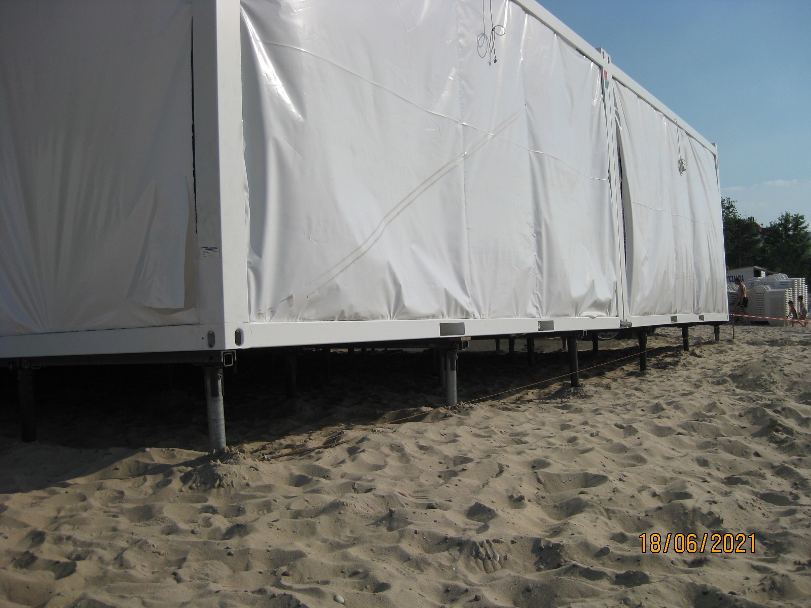 Mobilny fundament wkręcany w piasek pod obiekt kontenerowy z białym, plandekowym poszyciem, widoczne stalowe słupy i podkonstrukcja, montaż na plaży.