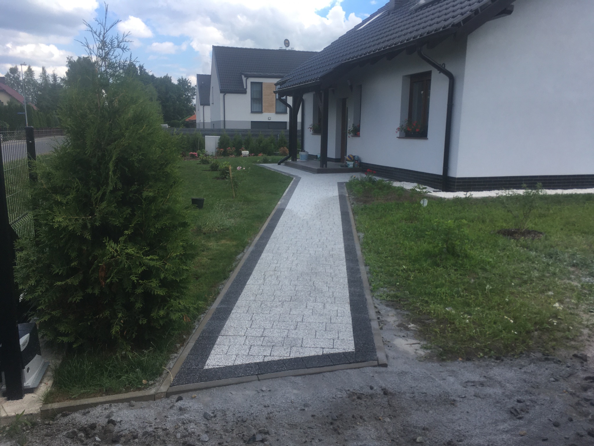 Nowoczesny, prosty chodnik z jasnoszarej kostki brukowej z ciemnoszarą obwódką prowadzący do domu z białymi ścianami i ciemnym dachem, widoczny trawnik i tuja.
