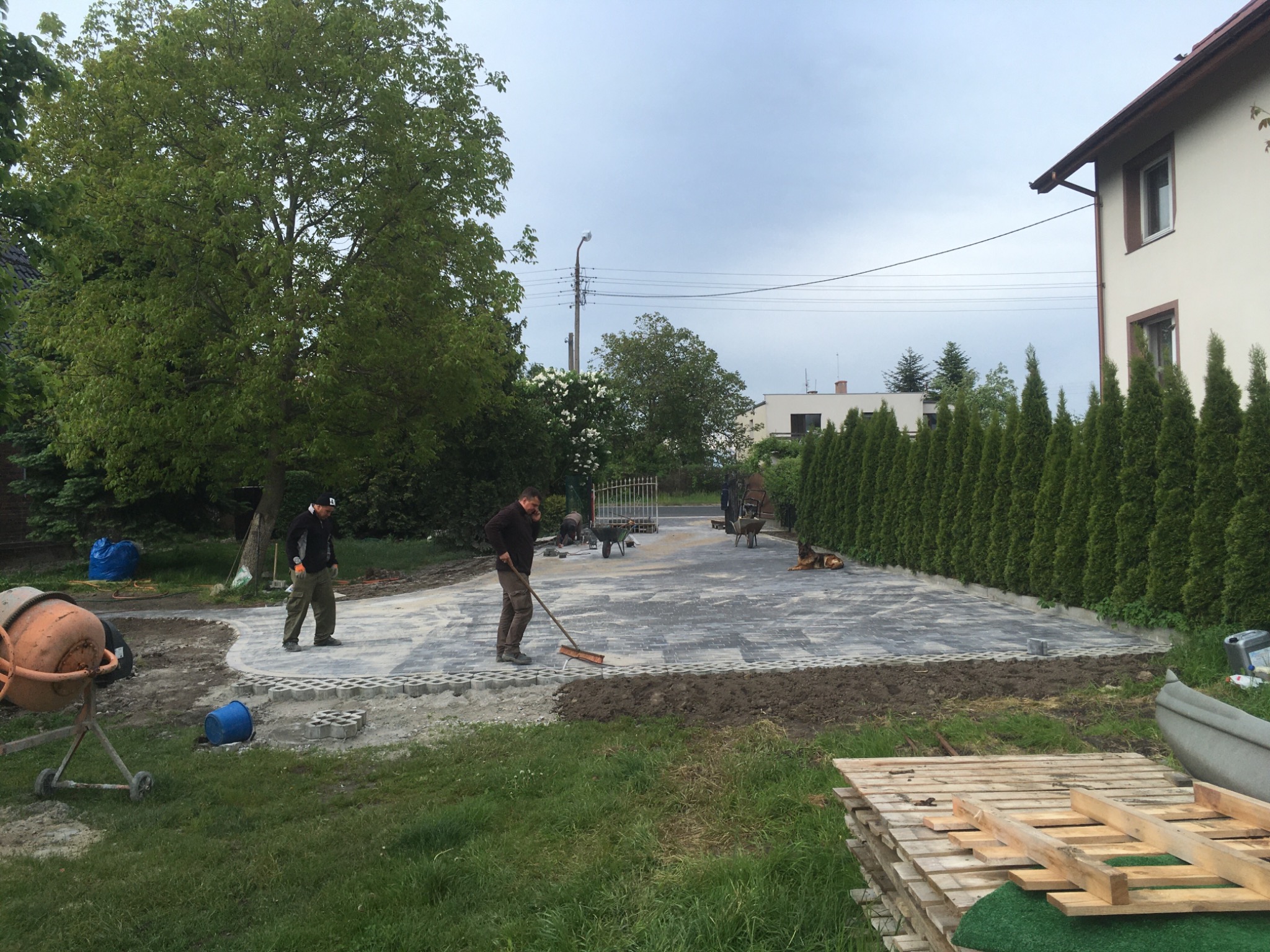 Prace brukarskie w toku: dwóch mężczyzn układa szarą kostkę na podjeździe, w tle betonowa mieszarka, ułożone palety i żywopłot z tui.