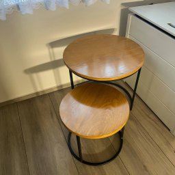 Krzysztof Orłowski - Dwa okrągłe stoliki kawowe z drewnianymi blatami i czarnymi metalowymi nogami ustawione jeden na drugim, obok białej komody i pod oknem z koronkową firanką.