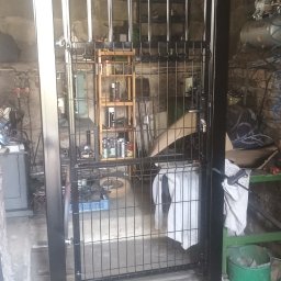 Krzysztof Orłowski - Czarna brama metalowa z prętami, w warsztacie, z widocznym regałem i sprzętem. Brama z łukiem u góry, w surowym, betonowym otoczeniu.