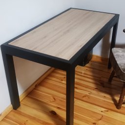 Krzysztof Orłowski - Stół z blatem imitującym drewno i czarnymi, metalowymi nogami. Minimalistyczny design, widoczna struktura drewna na blacie i solidne wykonanie.
