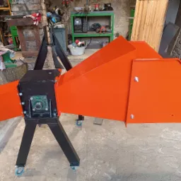 Pomarańczowo-czarna rębak do gałęzi na kółkach, z widocznymi śrubami i łożyskiem, stoi w warsztacie na betonowej podłodze. W tle narzędzia i ceglana ściana.