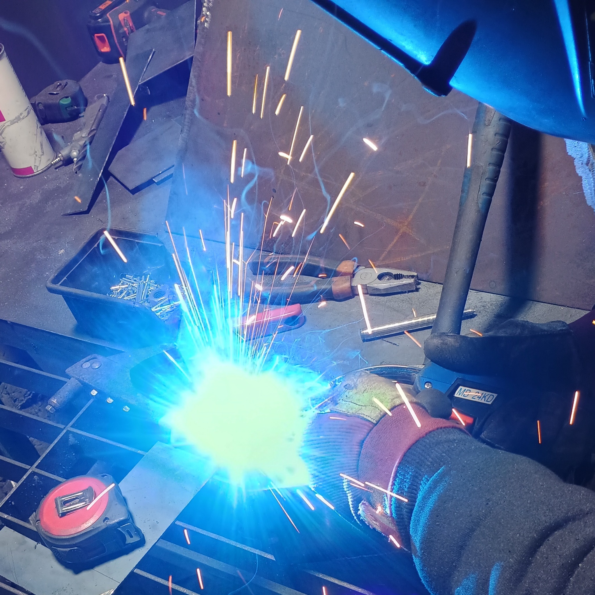 Spawanie metalu w warsztacie, widoczne iskry, spawarka w dłoni spawacza w rękawicy ochronnej, obok leżą narzędzia i pojemnik ze śrubami.