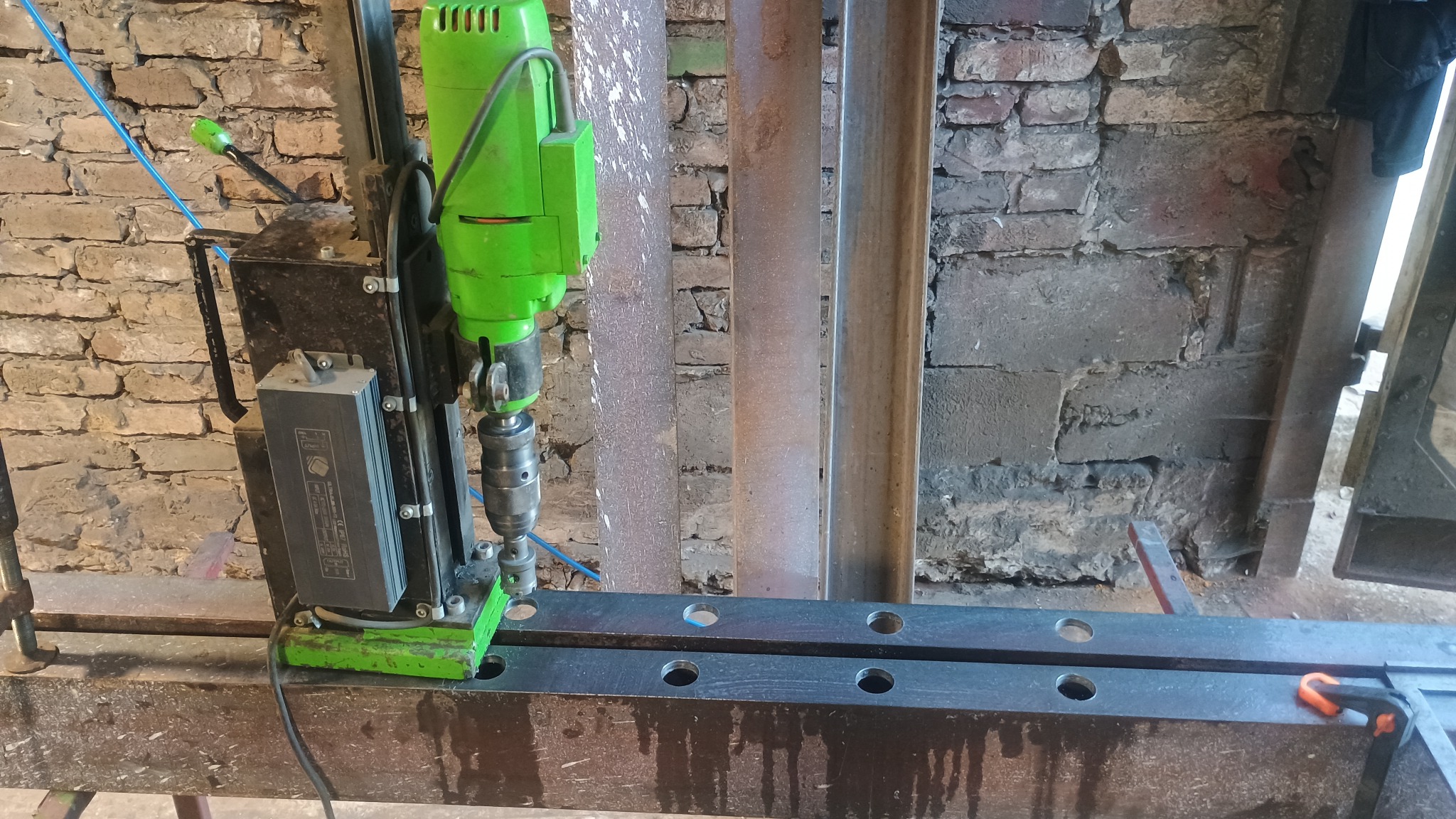 Wiertarka magnetyczna w kolorze zielonym wierci otwory w metalowym profilu na tle ściany z cegły