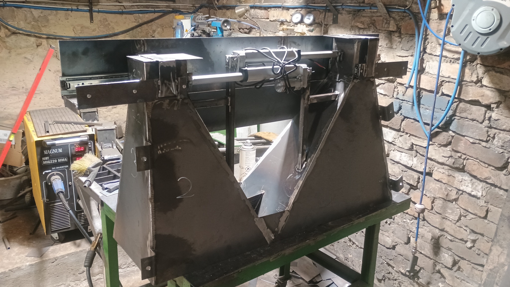 Konstrukcja z surowej stali z siłownikami pneumatycznymi, spawana w warsztacie, obok spawarka Magnum MIG220 MMA, na tle murowanej ściany.