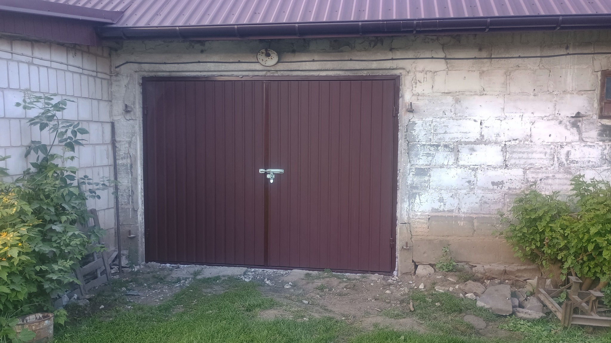 Brązowa, dwuskrzydłowa brama garażowa z metalu, z zamkiem na środku, zamontowana w murowanym garażu z widocznymi śladami użytkowania i roślinnością po bokach.