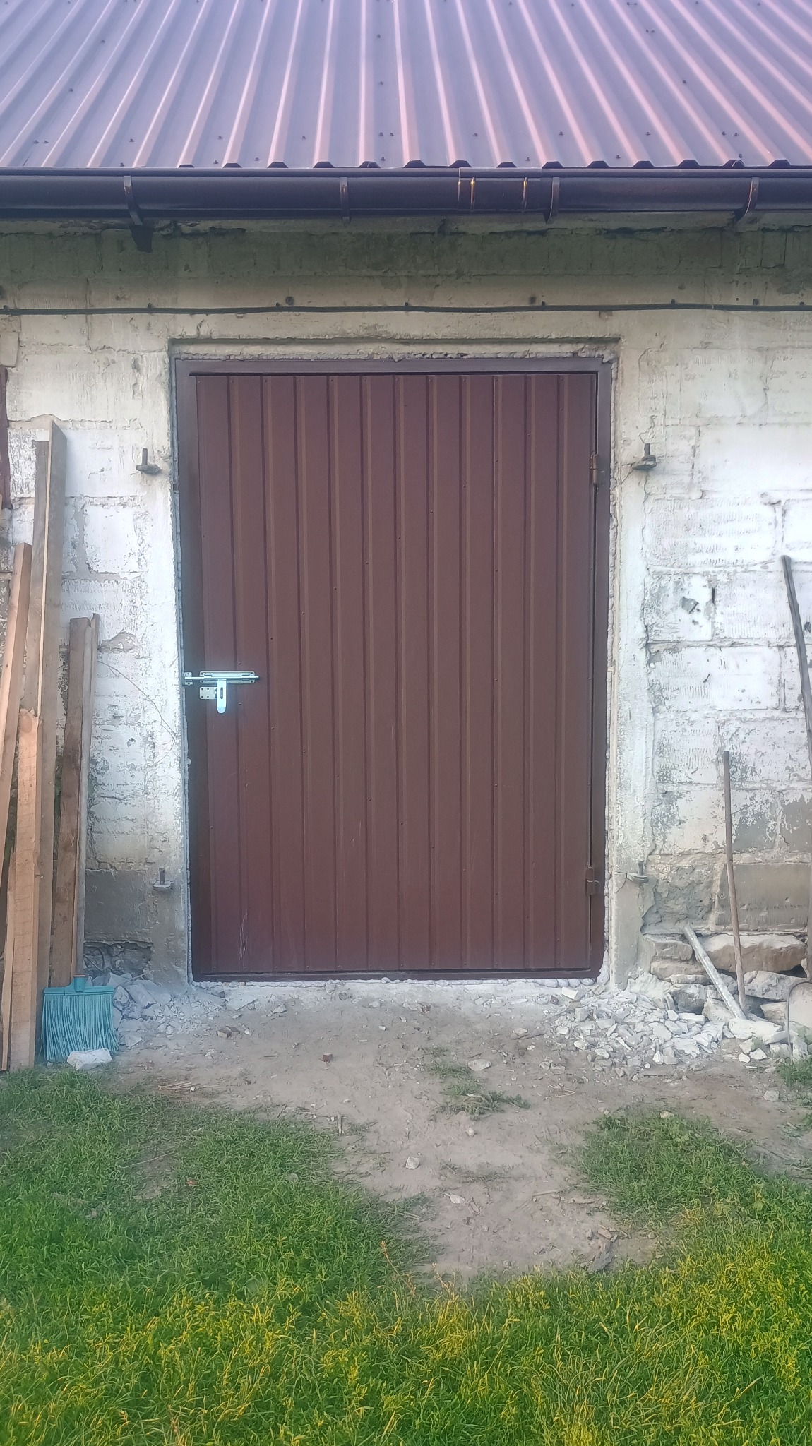 Brązowe, metalowe drzwi z zasuwką na tle starego, białego muru z betonu komórkowego i zielonej trawy. Widoczne deski i narzędzia.