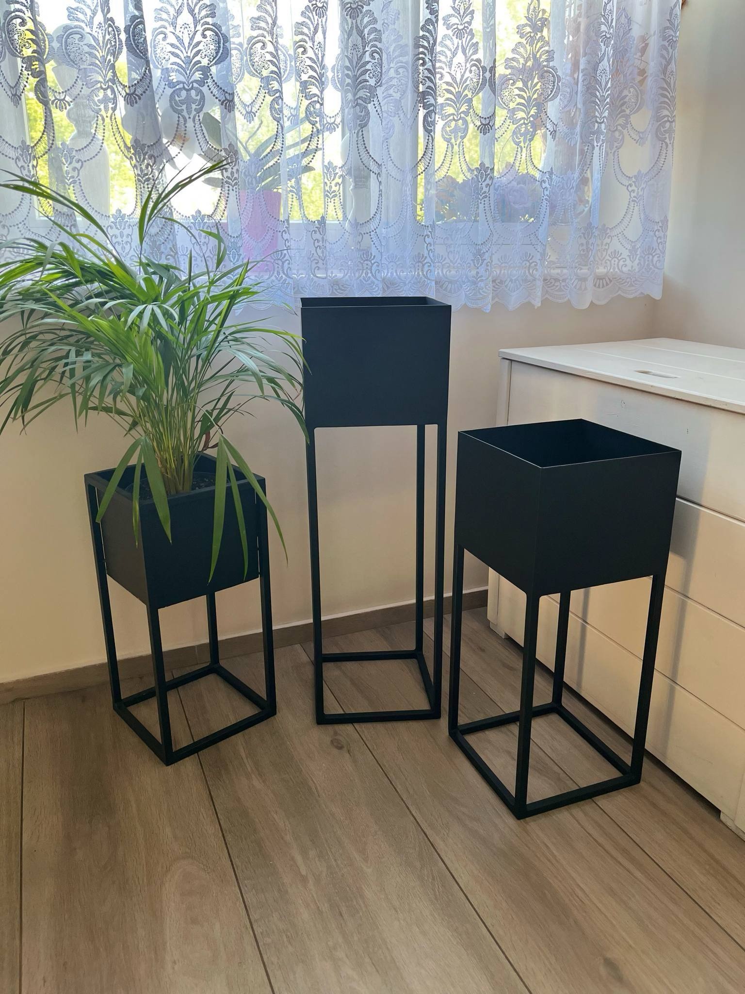 Trzy metalowe kwietniki w stylu loft, czarne, o różnych wysokościach, jeden z rośliną, na drewnianej podłodze, z firanką w tle. Minimalistyczny design.
