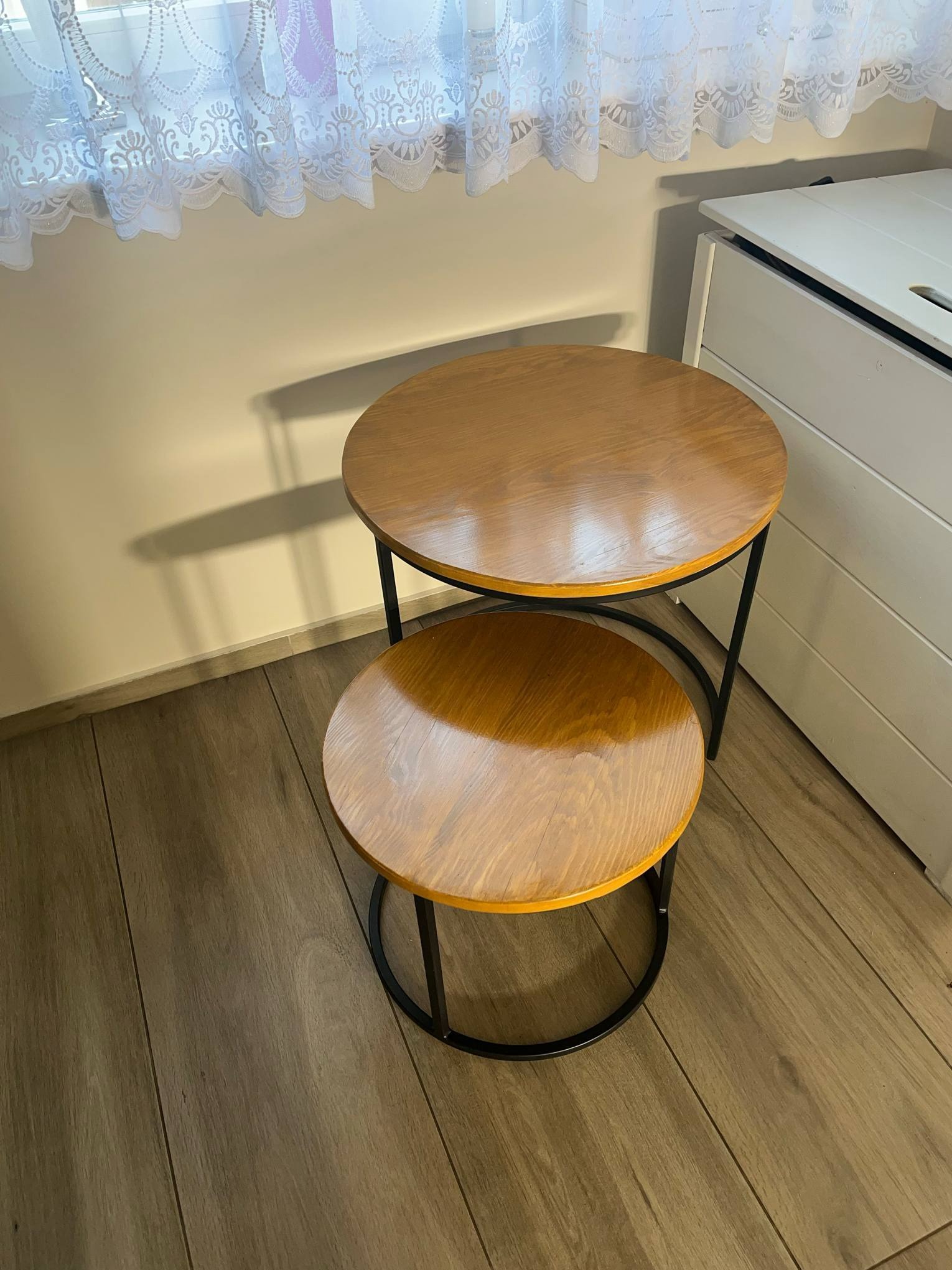 Dwa okrągłe stoliki kawowe z drewnianymi blatami i czarnymi metalowymi nogami ustawione jeden na drugim, obok białej komody i pod oknem z koronkową firanką.