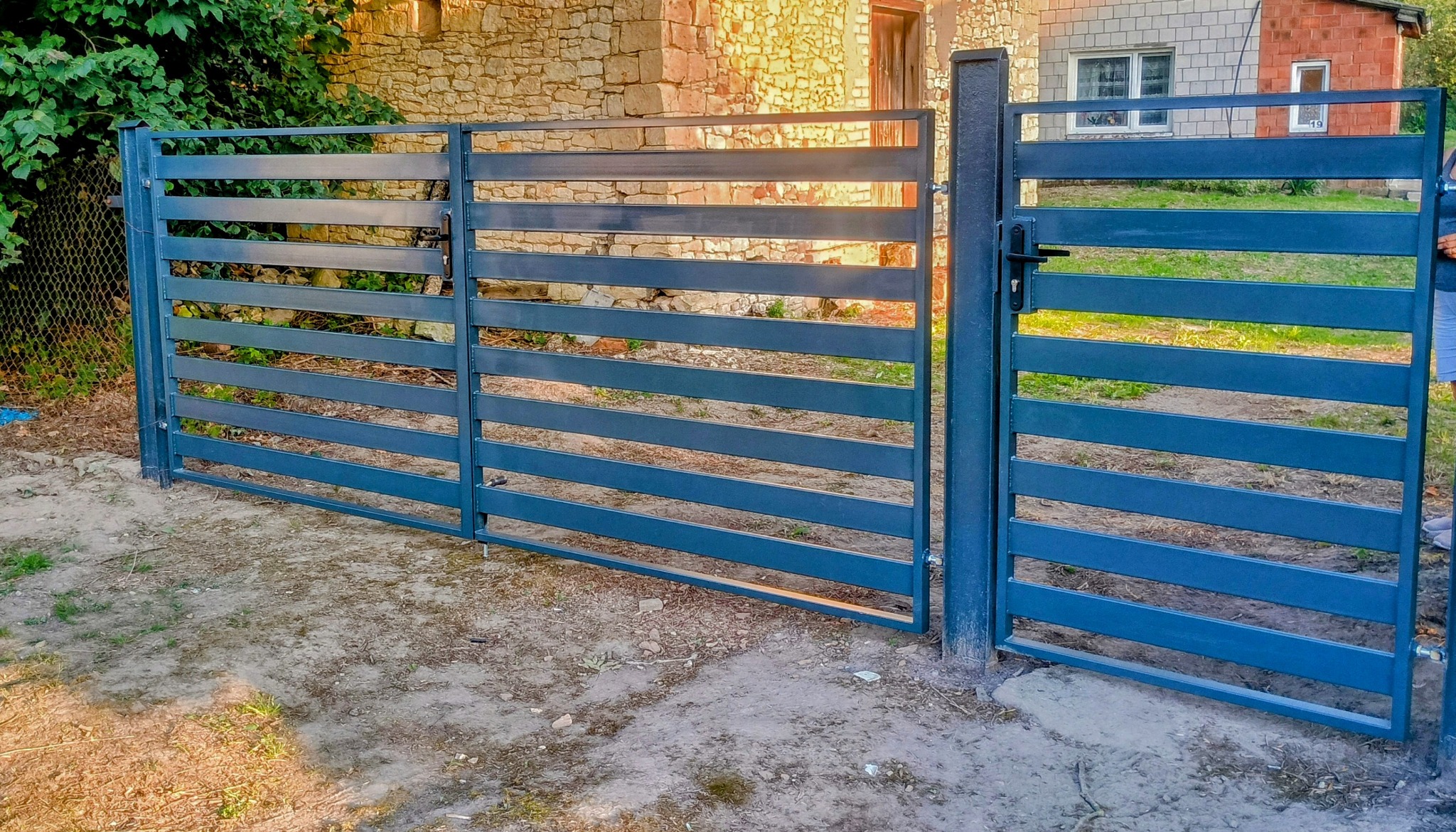 Nowoczesna, pozioma brama wjazdowa z metalu w kolorze grafitowym. Widoczne poziome profile i solidne słupki. Tło stanowi budynek z kamienia i zieleń.