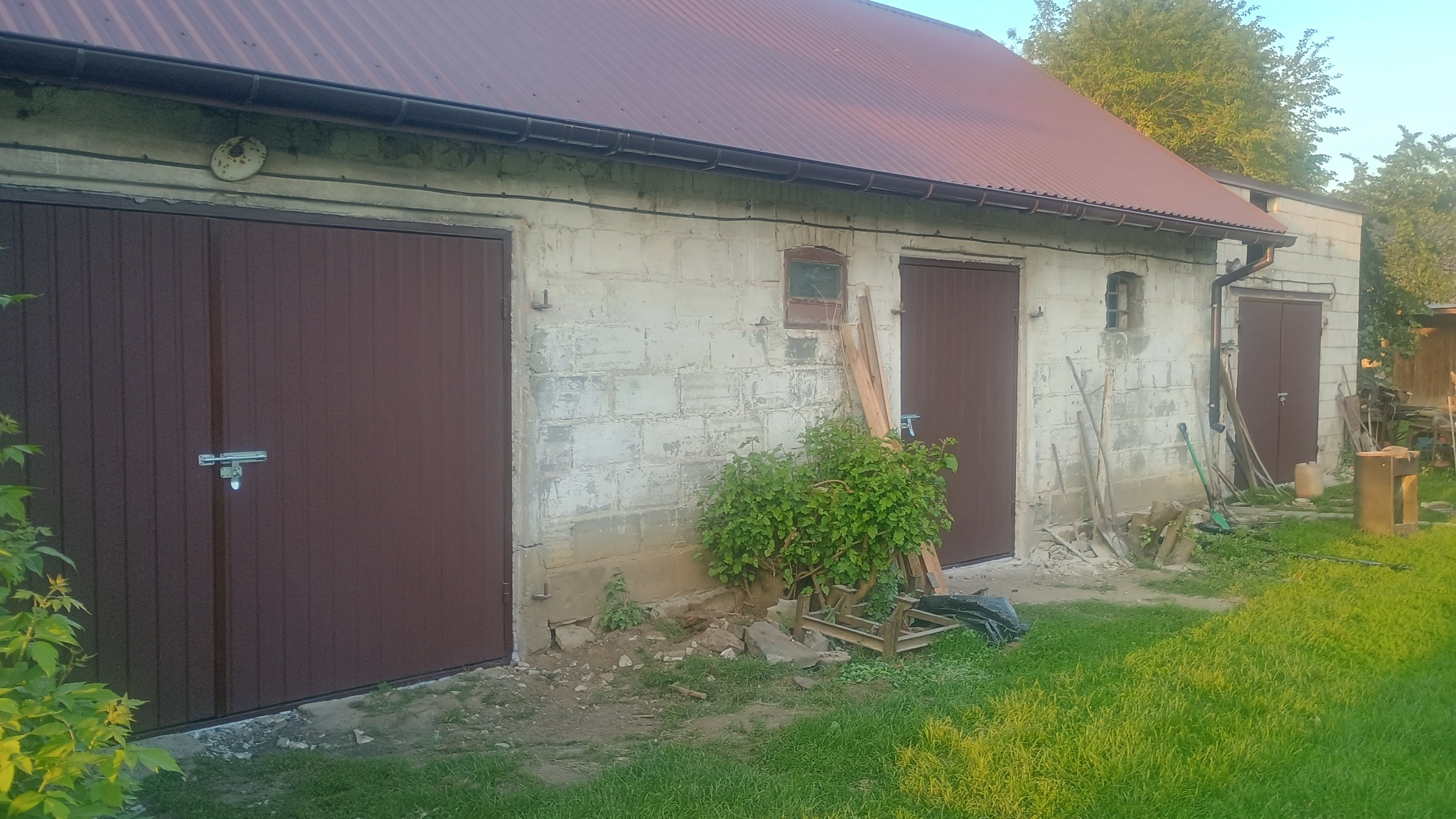 Brązowe, segmentowe bramy garażowe w budynku z szarego betonu, z widocznymi śladami użytkowania. Na dachu blacha trapezowa w kolorze ceglastym.