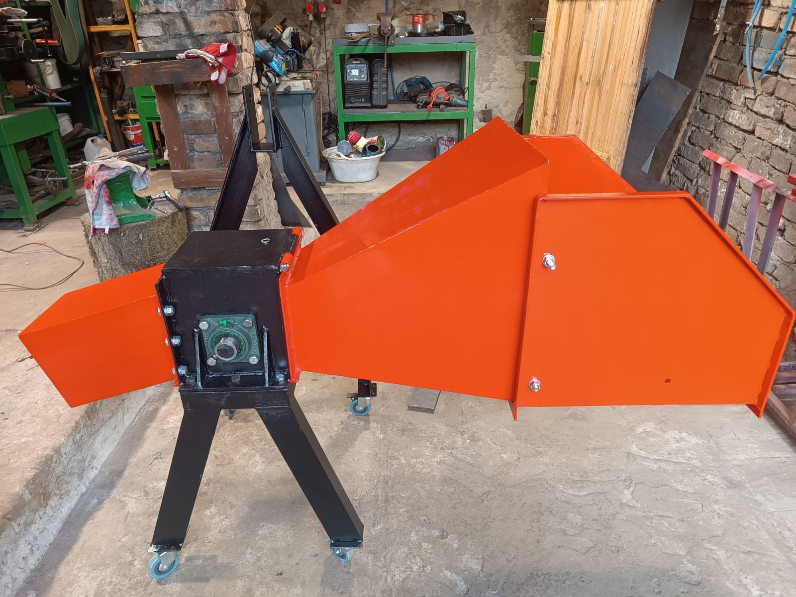 Pomarańczowo-czarna rębak do gałęzi na kółkach, z widocznymi śrubami i łożyskiem, stoi w warsztacie na betonowej podłodze. W tle narzędzia i ceglana ściana.