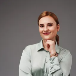 Portret uśmiechniętej kobiety z rudymi włosami, w satynowej koszuli i zegarku, opierającej podbródek na dłoni na szarym tle.