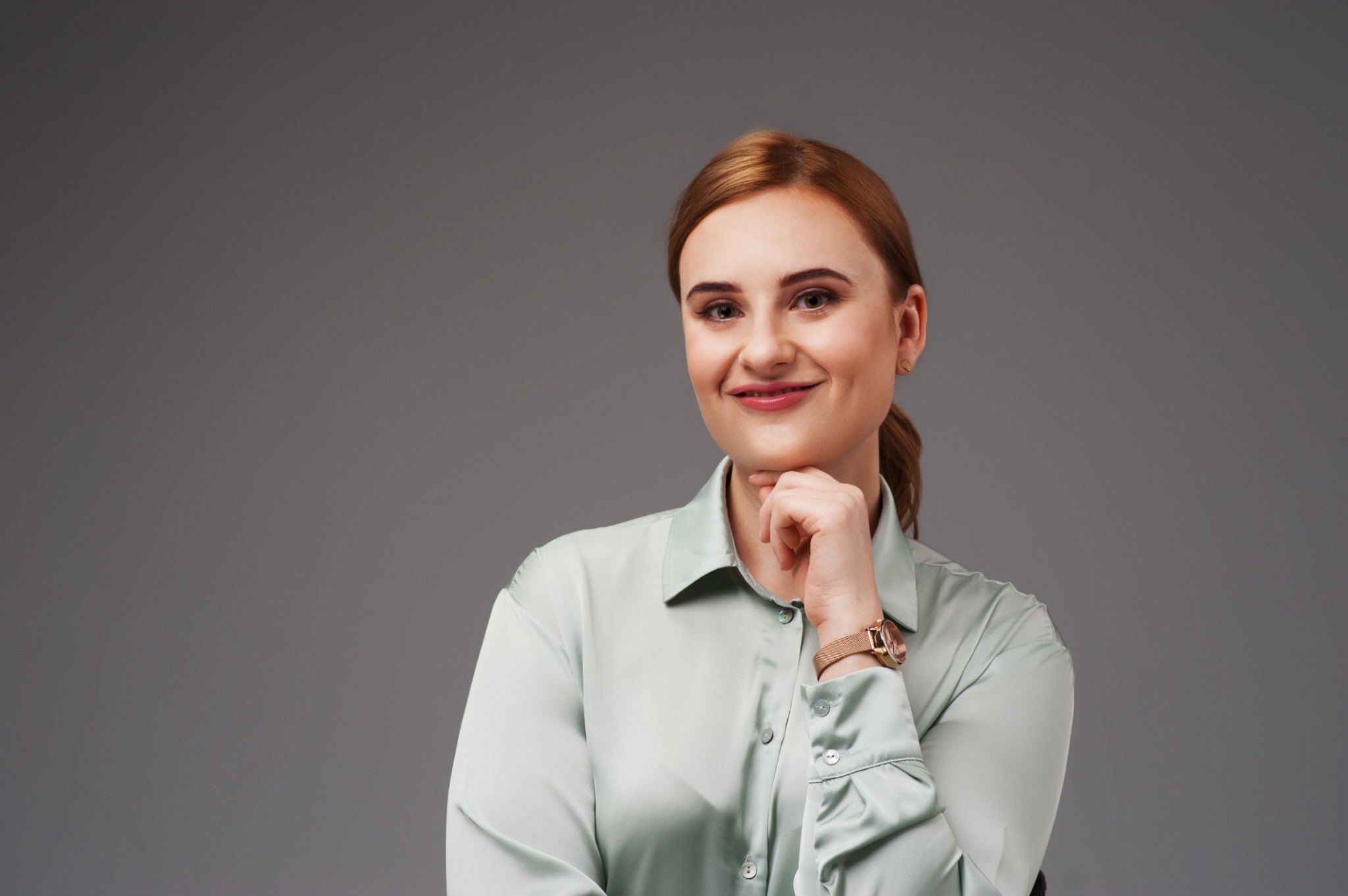 Portret uśmiechniętej kobiety z rudymi włosami, w satynowej koszuli i zegarku, opierającej podbródek na dłoni na szarym tle.