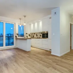 Jasne, przestronne wnętrze mieszkania po remoncie: drewnopodobne panele podłogowe, biała zabudowa kuchenna z geometrycznym backsplash, widok na miasto za dużymi oknami.