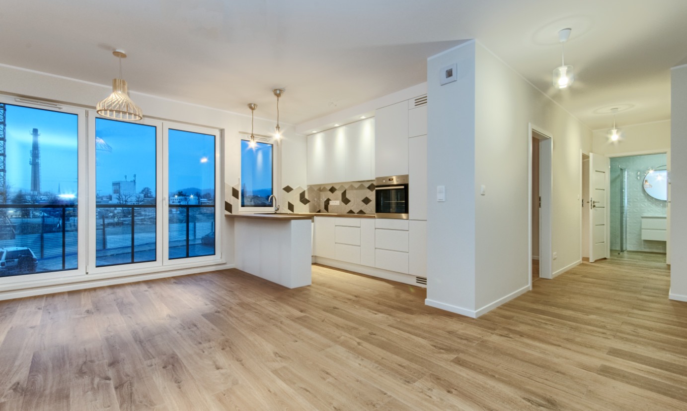 Jasne, przestronne wnętrze mieszkania po remoncie: drewnopodobne panele podłogowe, biała zabudowa kuchenna z geometrycznym backsplash, widok na miasto za dużymi oknami.