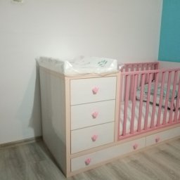 P. W. Mebel - Biało-różowe łóżeczko dziecięce z komodą i przewijakiem, ozdobione uchwytami w kształcie korony, stoi na drewnianej podłodze przy ścianie w dwóch kolorach.