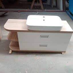 P. W. Mebel - Szafka łazienkowa z umywalką, białe fronty szuflad, drewniany blat z zaokrąglonymi krawędziami, widoczne półki z boku, całość ustawiona w warsztacie na podkładkach.