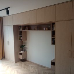 P. W. Mebel - Zabudowa meblowa w salonie z szafami, półkami i miejscem na telewizor, wykonana z jasnego drewna, z podłogą ułożoną w jodełkę i oświetleniem szynowym na suficie.