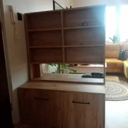 Drewniany regał z szafkami na dole, widoczne półki i uchwyty, w tle sofa i dywan.