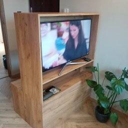 P. W. Mebel - Drewniana zabudowa telewizora z półkami, w odcieniach naturalnego drewna, z widocznym telewizorem i rośliną doniczkową po prawej stronie.