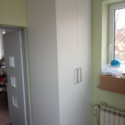 P. W. Mebel - Biała, wysoka szafa z dwoma drzwiami z metalowymi uchwytami, stojąca w rogu pokoju z widocznym oknem i grzejnikiem, obok otwartych drzwi do innego pomieszczenia.