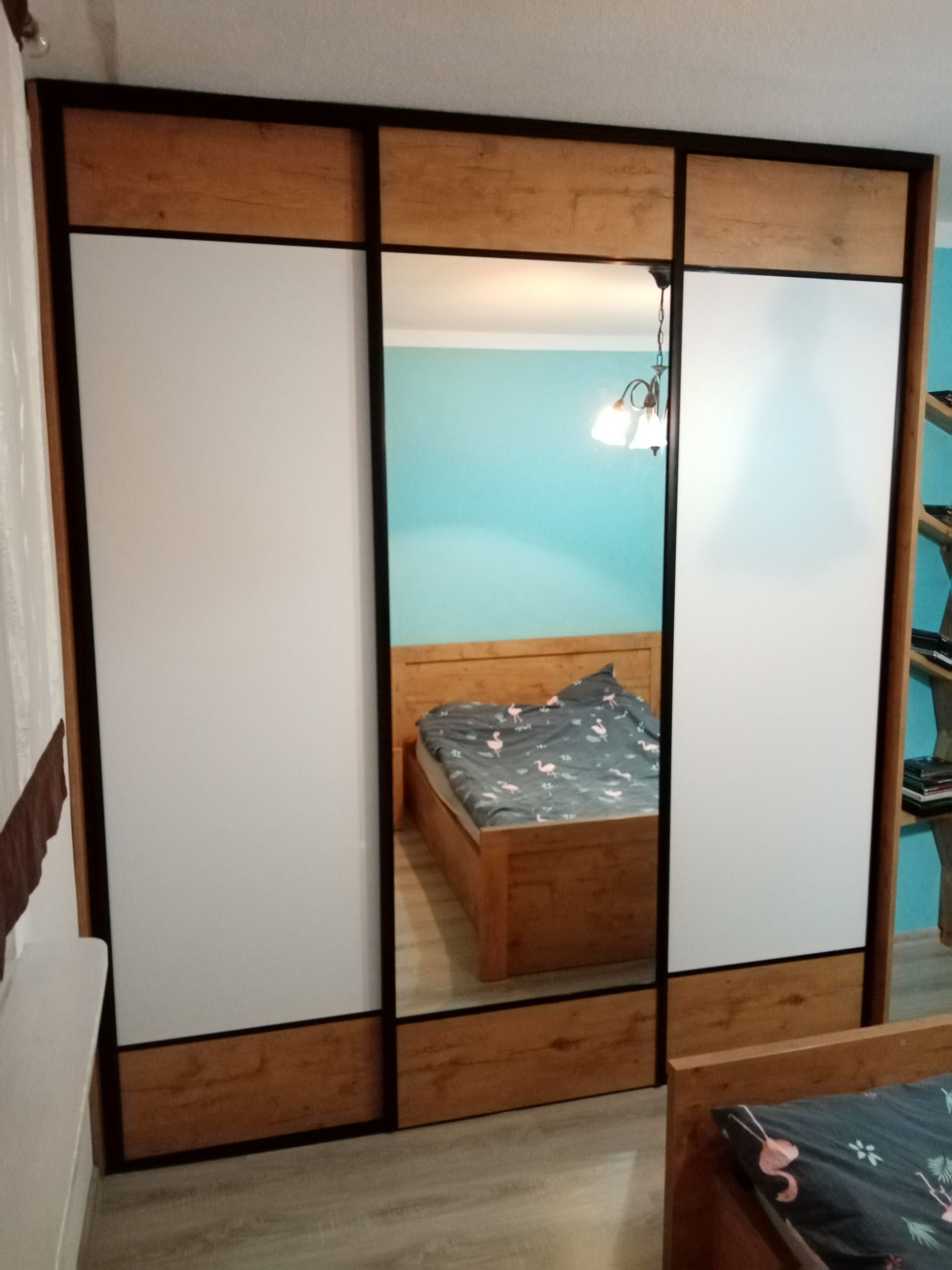 Trzydrzwiowa szafa wnękowa z przesuwnymi drzwiami: białe panele, drewnopodobne wstawki i lustrzane odbicie sypialni z łóżkiem, lampą i turkusową ścianą.