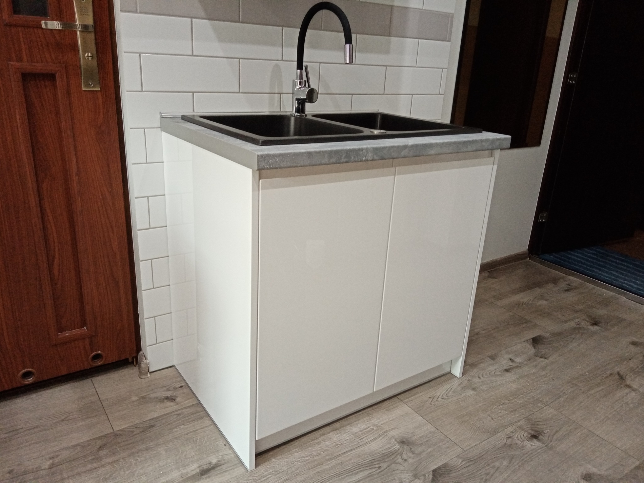 Biała szafka pod umywalkę z szarym blatem i czarną baterią, na tle białych płytek i drewnianej podłogi oraz fragmentu drzwi.