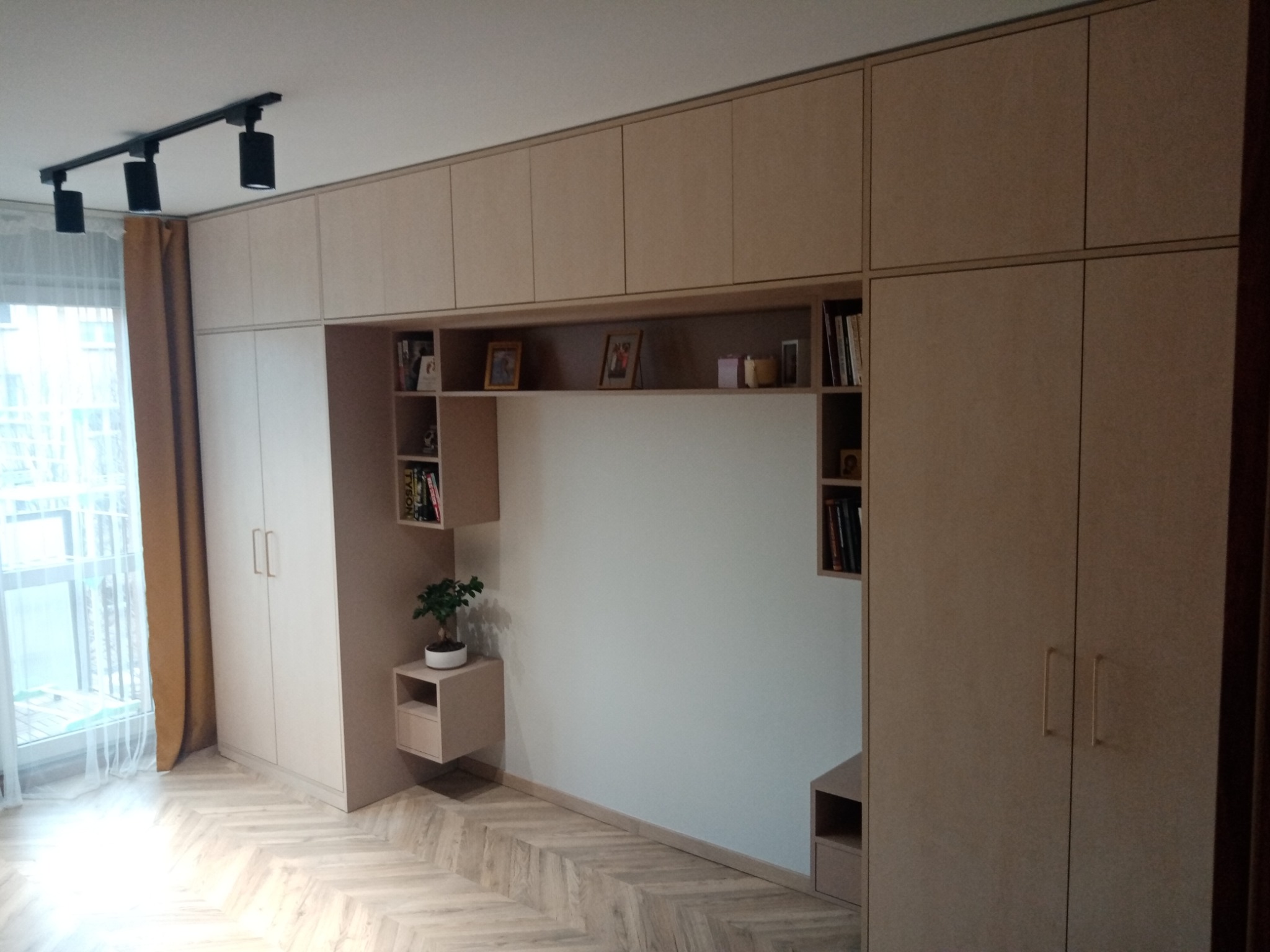 Zabudowa meblowa w salonie z szafami, półkami i miejscem na telewizor, wykonana z jasnego drewna, z podłogą ułożoną w jodełkę i oświetleniem szynowym na suficie.