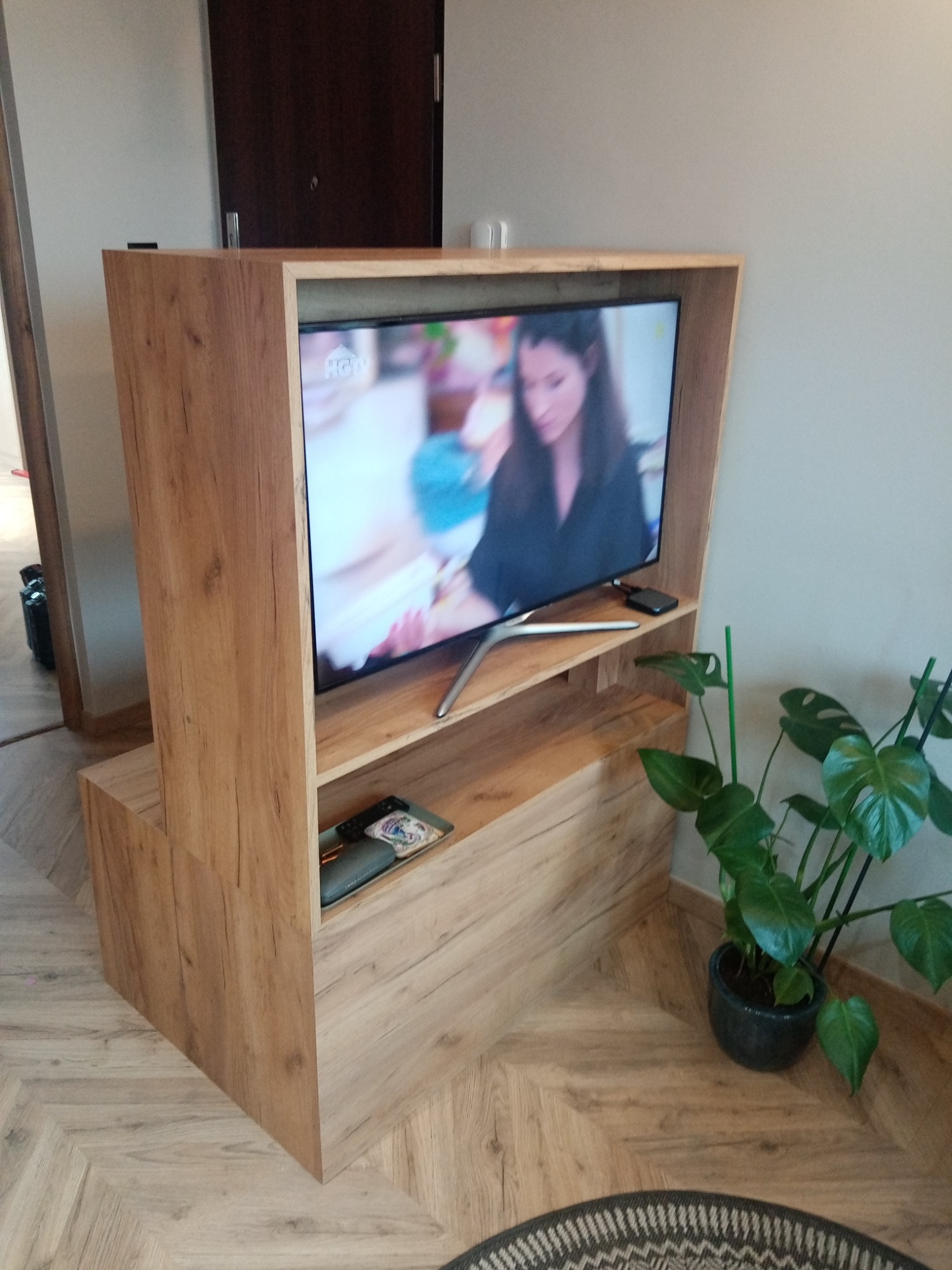 Drewniana zabudowa telewizora z półkami, w odcieniach naturalnego drewna, z widocznym telewizorem i rośliną doniczkową po prawej stronie.