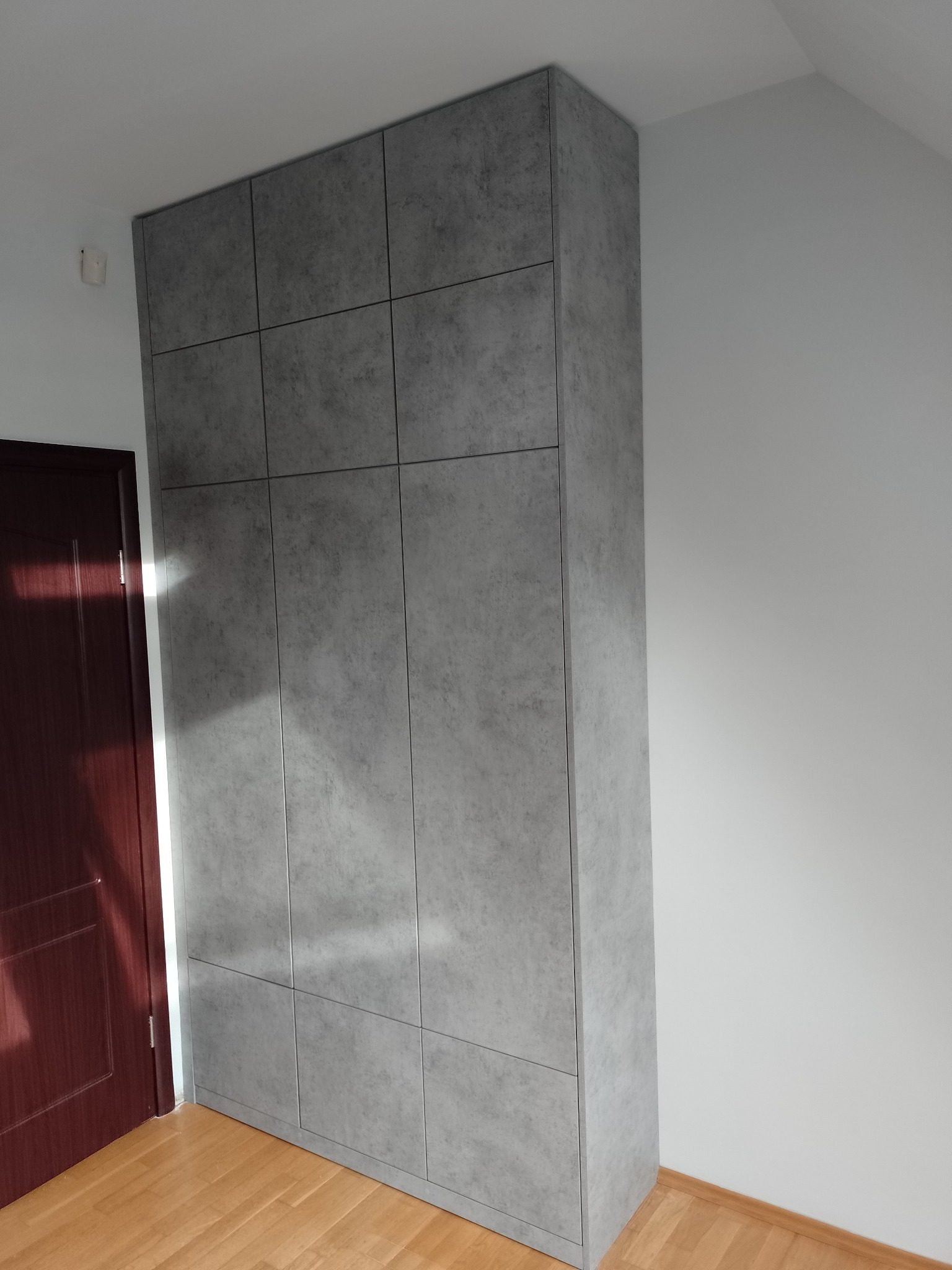 Wysoka, szara szafa wnękowa z panelami imitującymi beton, umieszczona w rogu pokoju z drewnianą podłogą i brązowymi drzwiami.