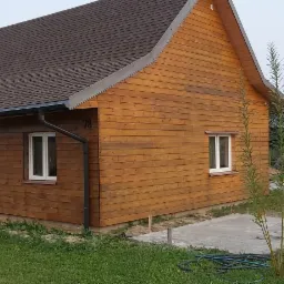 Nowo wybudowany, drewniany dom z brązowym dachem, widoczne rynny i numer 78 na elewacji bocznej. Obok domu świeżo wylany betonowy fundament i zwinięty wąż ogrodowy.