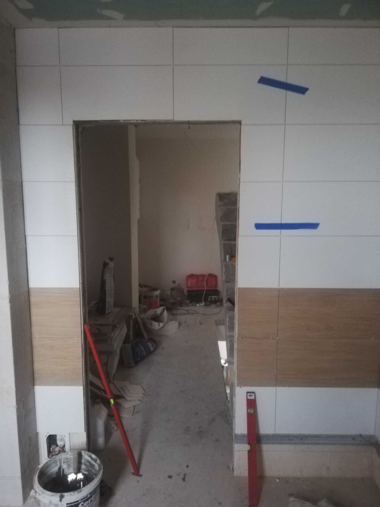 Wnętrze w trakcie remontu łazienki: częściowo wyłożone białymi i drewnopodobnymi płytkami, z niebieską taśmą malarską na górnych płytkach, widoczne narzędzia i materiały budowlane w tle, poziomica...