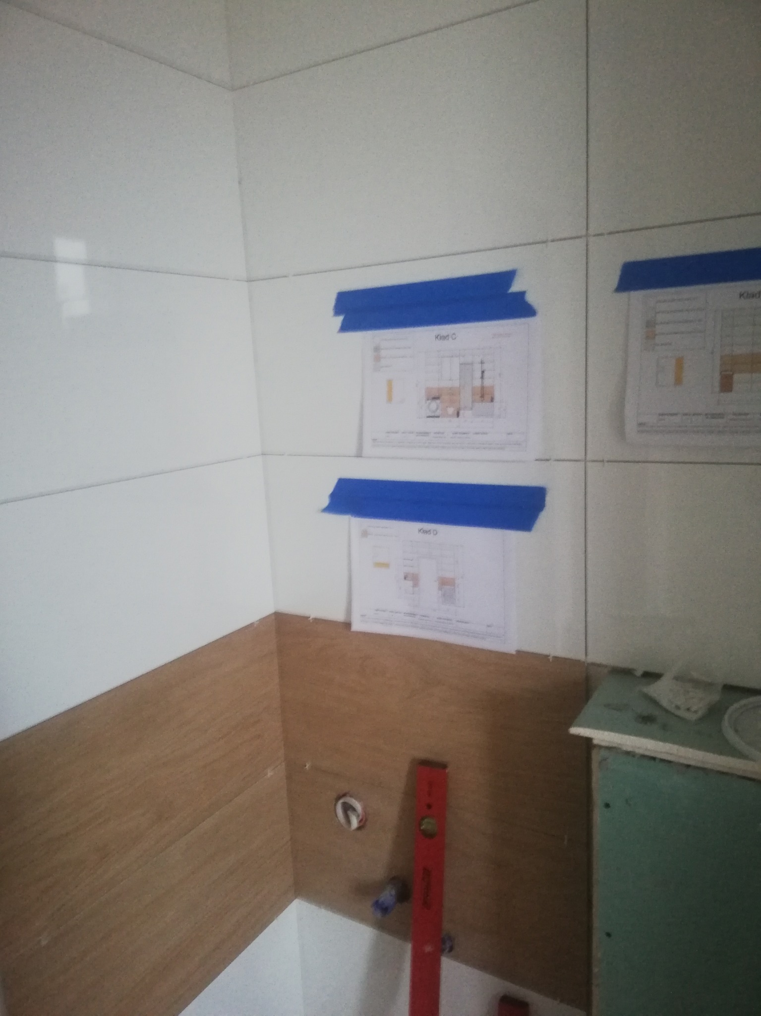 Łazienka w trakcie wykańczania: białe płytki ceramiczne na ścianach, częściowo wykończone drewnopodobną okładziną, z planami technicznymi przyklejonymi niebieską taśmą i czerwoną poziomicą.