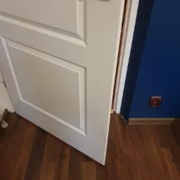 Otwarta biała płycinowa drzwi z metalową klamką na tle ciemnoniebieskiej ściany i brązowej drewnianej podłogi.